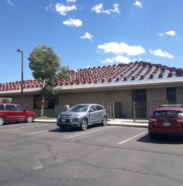 A building with a red-tile roof and multiple parked cars under a clear blue sky, featuring a flagpole on the rooftop.