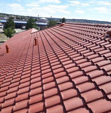 A sloped red tile roof featuring several chimney pipes, with a distant view of buildings and trees under a blue sky.