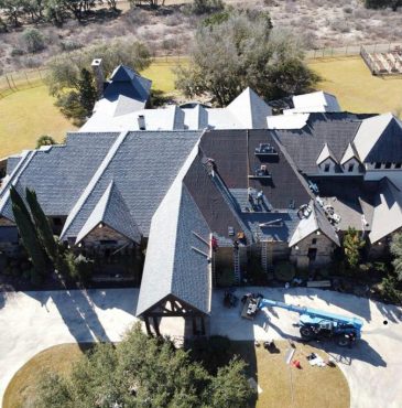 Aerial view of a large stone house with ongoing roof renovations, equipment, and workers visible against a scenic landscape.